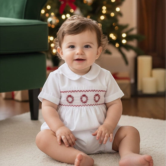 Carriage Boutique white batiste boys shortall with red hand-smocked holiday wreath detail, worn by a baby boy.