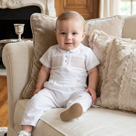 Carriage Boutique white cotton boys christening outfit with hand-smocked detail and satin trim worn by a baby on a sofa