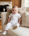 Carriage Boutique white cotton boys christening outfit with hand-smocked detail and satin trim worn by a baby on a sofa