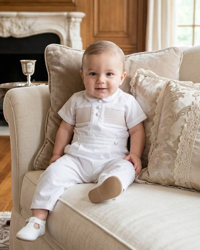 Carriage Boutique white cotton boys christening outfit with hand-smocked detail and satin trim worn by a baby on a sofa