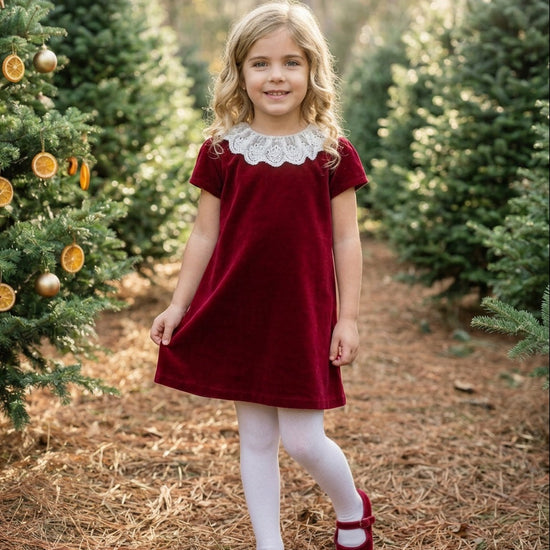 Carriage Boutique toddler girl wearing a burgundy velvet dress with a white lace collar in a festive forest setting.