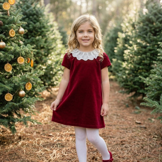 Young girl wearing a Carriage Boutique burgundy velvet dress with a white lace collar in a festive forest setting.