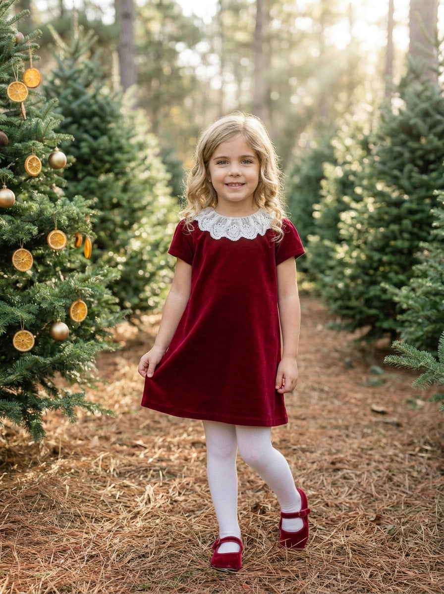 Young girl wearing a Carriage Boutique burgundy velvet dress with a white lace collar in a festive forest setting.