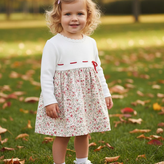 Carriage Boutique baby girl wearing a white knit long sleeve dress with a floral skirt and red ribbon detail in a park