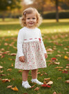 Carriage Boutique baby girl wearing a white knit long sleeve dress with a floral skirt and red ribbon detail in a park
