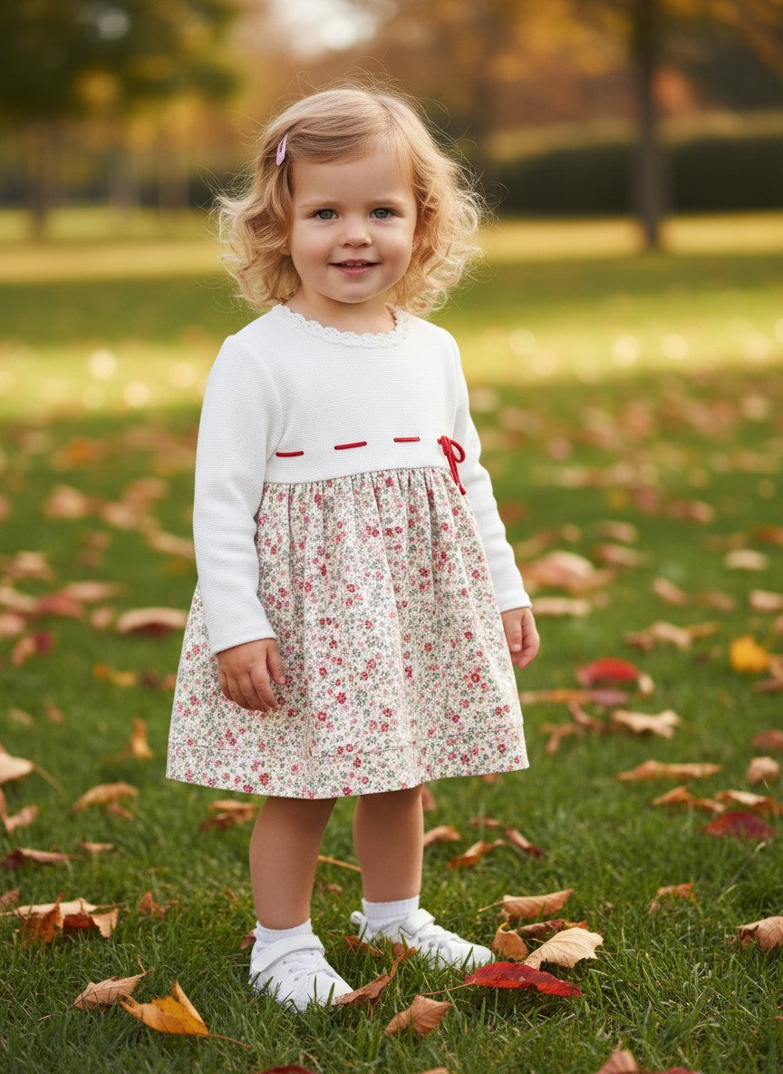 Carriage Boutique baby girl wearing a white knit long sleeve dress with a floral skirt and red ribbon detail in a park