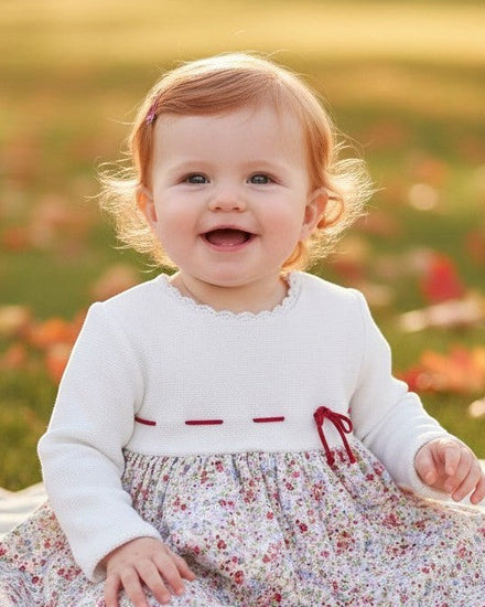 Carriage Boutique baby girl wearing a long sleeve white knit bodice dress with a floral skirt and red ribbon detail.