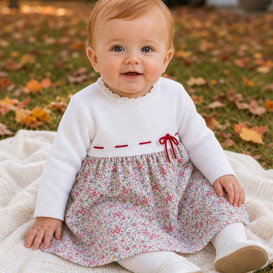 Carriage Boutique baby girl wearing a white knit long sleeve dress with a floral skirt and red ribbon waist detail.