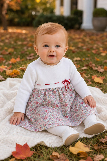 Carriage Boutique baby girl wearing a white knit long sleeve dress with a floral skirt and red ribbon waist detail.
