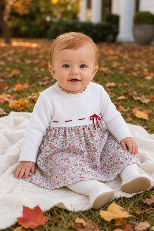 Carriage Boutique baby girl wearing a white knit long sleeve dress with a floral skirt and red ribbon waist detail.