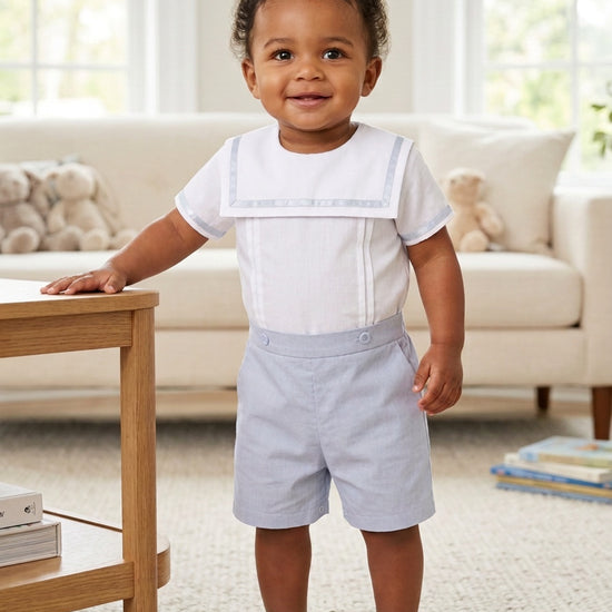 Carriage Boutique toddler boy wearing a white sailor-style shirt and light gray shorts in a bright living room.
