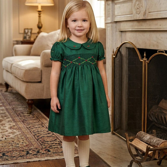 Carriage Boutique green silk toddler dress with smocked chest, Peter Pan collar, and puffy sleeves by a fireplace.