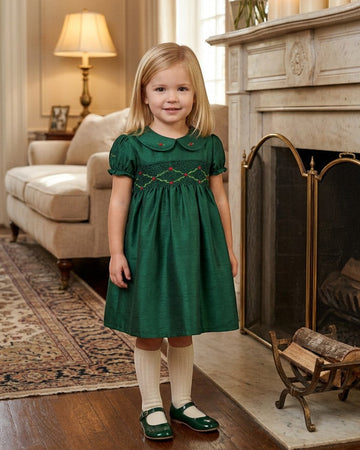 Carriage Boutique green silk toddler dress with smocked chest, Peter Pan collar, and puffy sleeves by a fireplace.