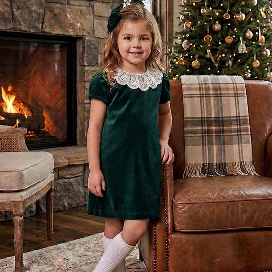 Carriage Boutique toddler girl in a green velvet dress with a white lace collar standing by a Christmas tree
