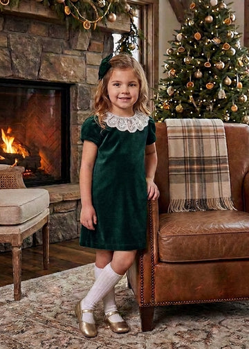 Carriage Boutique toddler girl in a green velvet dress with a white lace collar standing by a Christmas tree