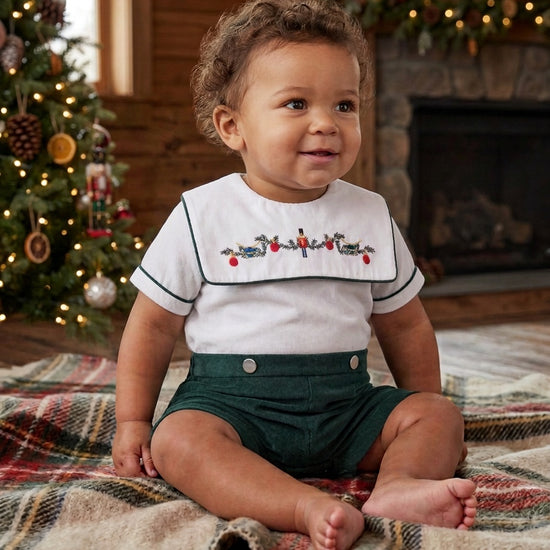 Carriage Boutique infant boy wearing a green and white smocked bobbie suit with a wooden soldier motif on a blanket.