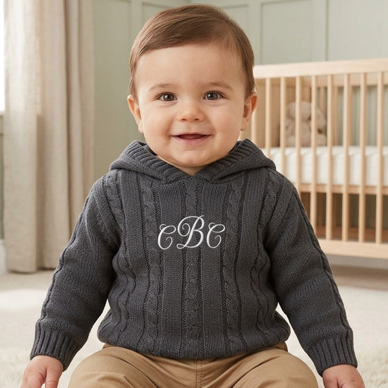 Carriage Boutique baby boy wearing a dark grey cable-knit hooded sweater with white monogram embroidery on the chest.