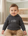 Carriage Boutique baby boy wearing a dark grey cable-knit hooded sweater with white monogram embroidery on the chest.