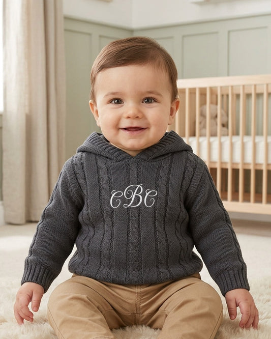 Carriage Boutique baby boy wearing a dark grey cable-knit hooded sweater with white monogram embroidery on the chest.