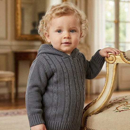 Toddler boy wearing a dark grey Carriage Boutique cable-knit hooded sweater with ribbed cuffs in a classic room.