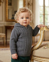 Toddler boy wearing a dark grey Carriage Boutique cable-knit hooded sweater with ribbed cuffs in a classic room.