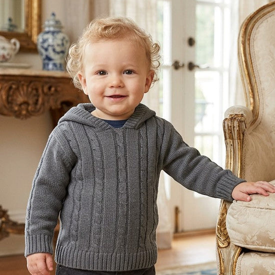 Carriage Boutique baby boy wearing a dark grey cable-knit hooded sweater with ribbed cuffs in a classic room setting.