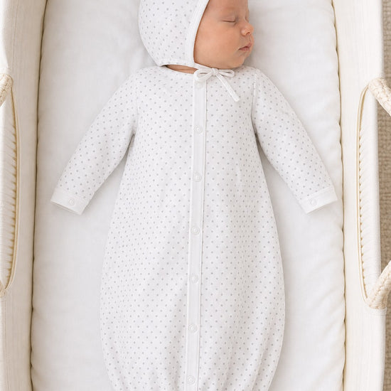 Newborn sleeping in a Carriage Boutique white gown and bonnet set with a grey star print in a bassinet.
