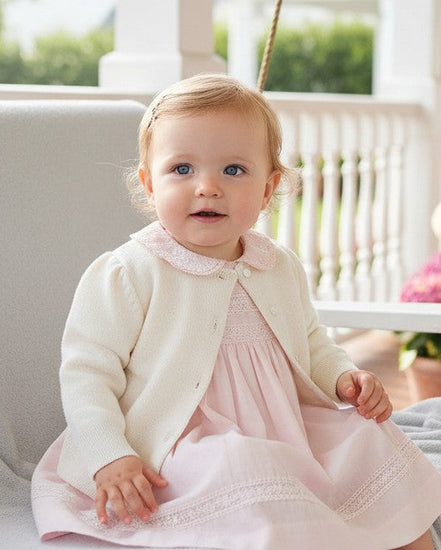 Carriage Boutique ivory cotton cashmere cardigan worn by a baby girl on a porch swing with a pink dress