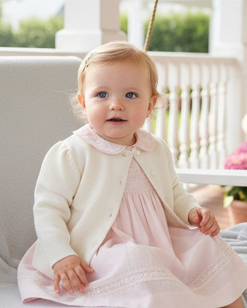 Carriage Boutique ivory cotton cashmere cardigan worn by a baby girl on a porch swing with a pink dress