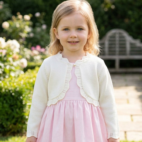 Carriage Boutique toddler girl wearing an ivory cotton bolero shrug with scalloped edges over a light pink dress