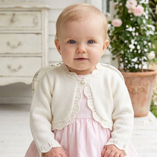 Carriage Boutique ivory cotton bolero shrug with delicate scalloped edges worn by a baby girl over a pink dress.