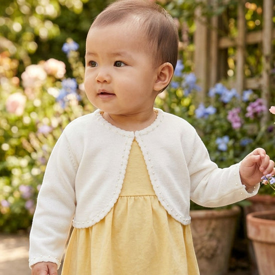 Carriage Boutique ivory knit bolero sweater with delicate pearl trim worn by a baby girl in a garden setting