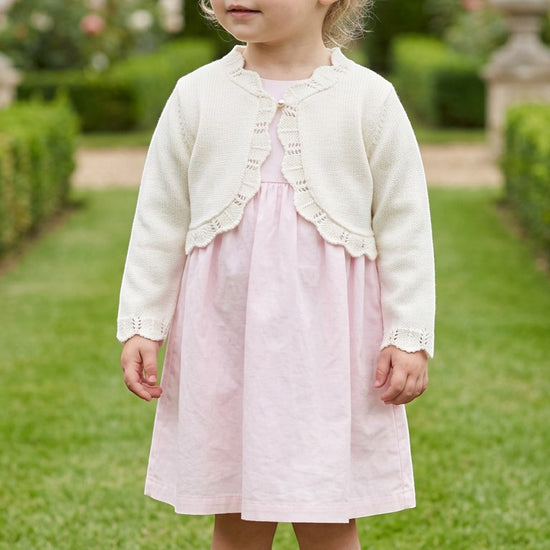 Carriage Boutique toddler girl wearing an ivory cotton bolero shrug with delicate scalloped edges over a pink dress