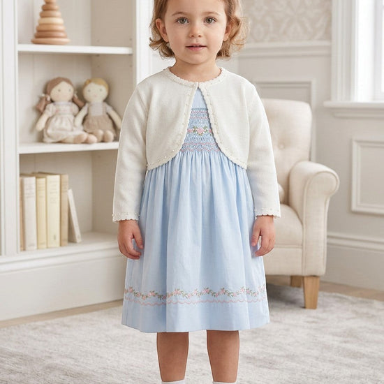 Toddler girl wearing a white Julius Berger bolero sweater over a light blue smocked dress in a bright nursery room.