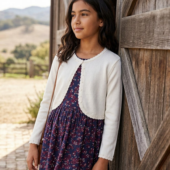 Carriage Boutique Julius Berger white bolero sweater worn by a young girl over a floral dress by a rustic barn door.