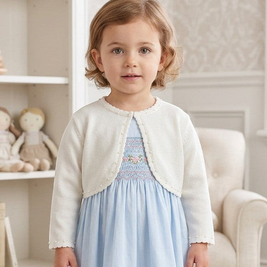 Carriage Boutique toddler girl wearing a white Julius Berger bolero sweater over a light blue smocked dress.
