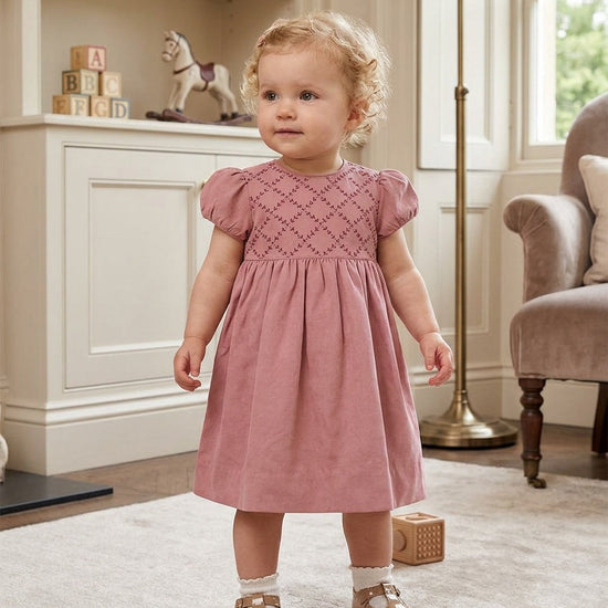Toddler girl wearing a Carriage Boutique mauve suede-like dress with a smocked, embroidered yoke and short sleeves.