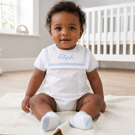 Carriage Boutique baby boy wearing a white bubble romper with a blue monogrammed collar, sitting on a knit blanket.