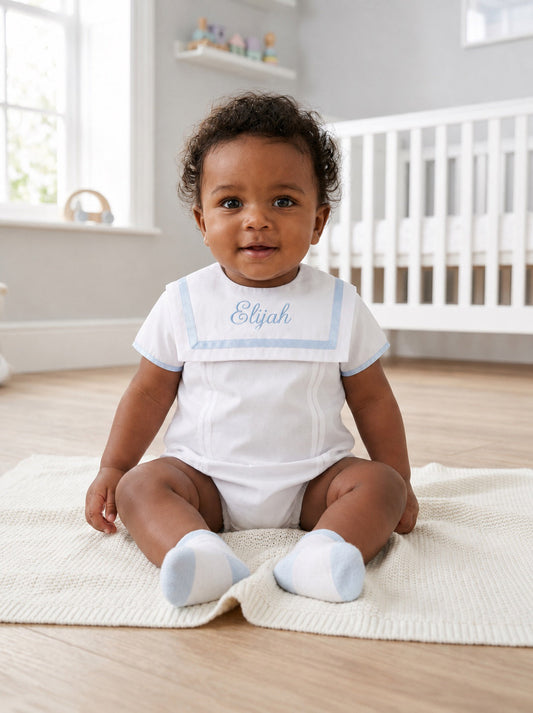 Carriage Boutique baby boy wearing a white bubble romper with a blue monogrammed collar, sitting on a knit blanket.