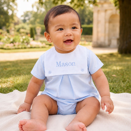 Carriage Boutique baby boy wearing a light blue cotton bubble romper with a white monogrammed bib sitting on a blanket