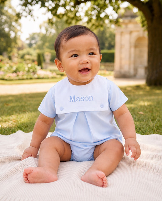 Carriage Boutique baby boy wearing a light blue cotton bubble romper with a white monogrammed bib sitting on a blanket