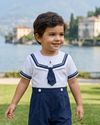 A young boy wearing a Carriage Boutique white and navy blue nautical bobby suit with a sailor collar and embroidery.