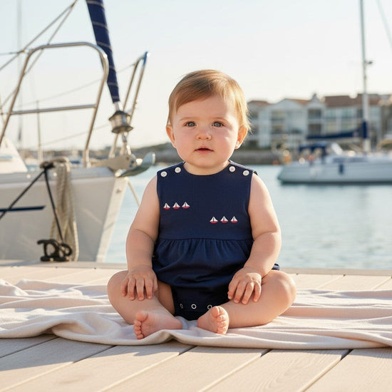 Carriage Boutique navy blue sleeveless baby romper with embroidered sailboats and red & white sails. Features shoulder buttons and snap closure.