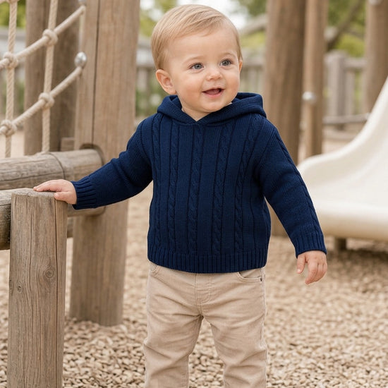Toddler boy wearing a navy cable knit hooded sweater from Carriage Boutique while standing on a playground