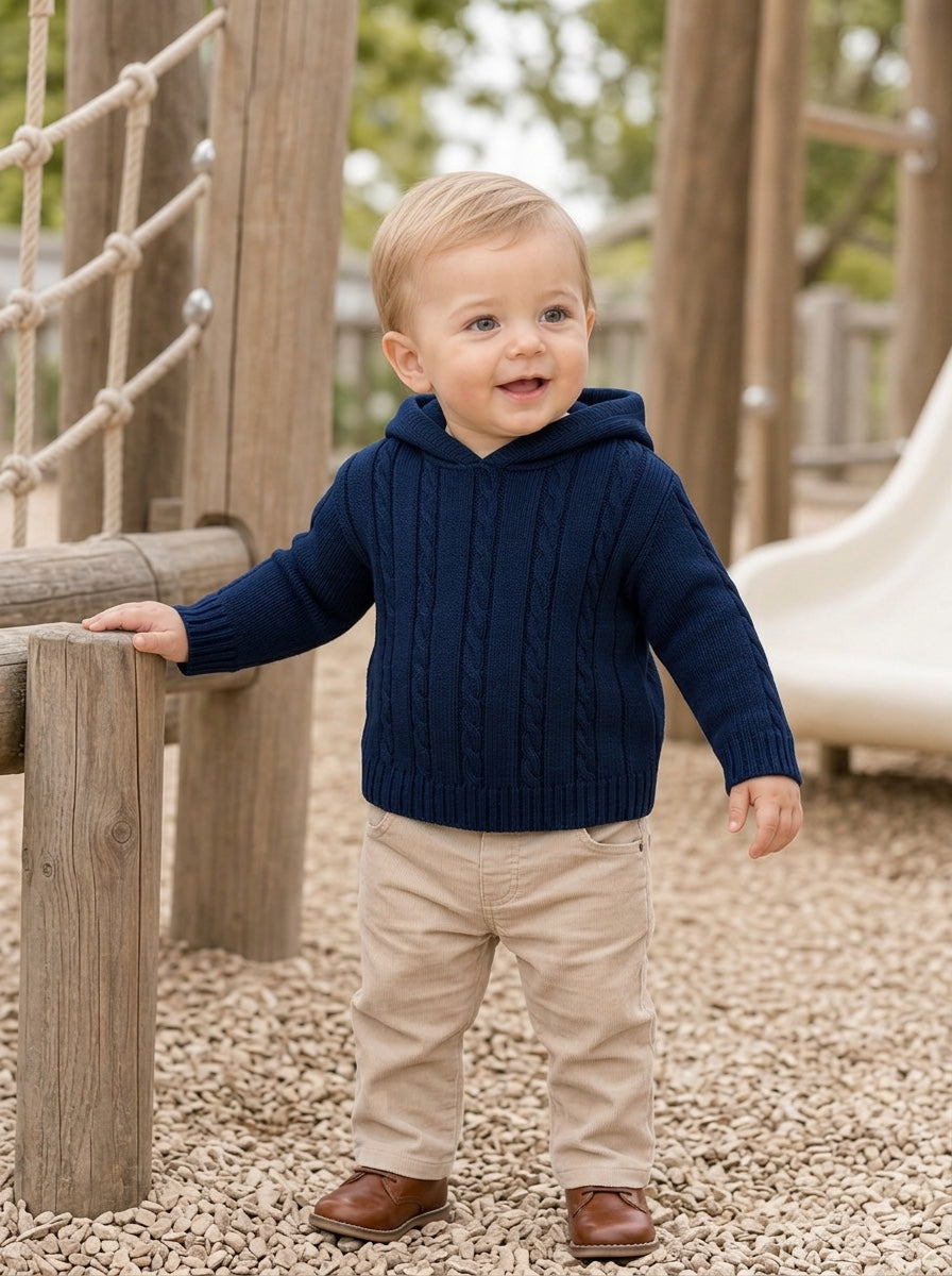 Toddler boy wearing a navy cable knit hooded sweater from Carriage Boutique while standing on a playground