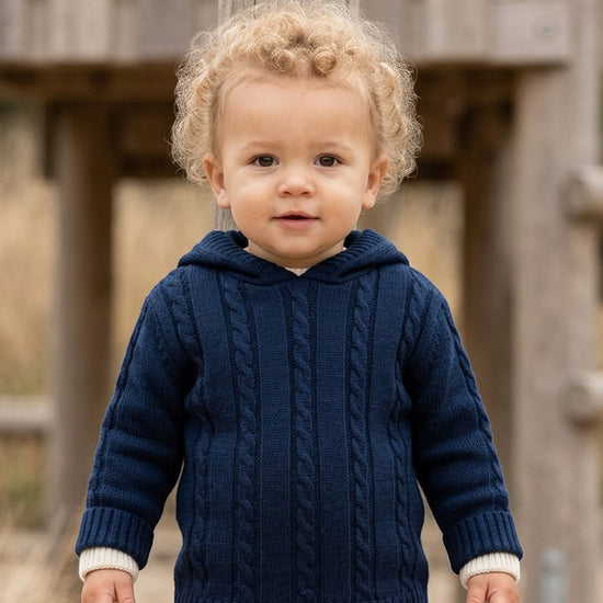 Carriage Boutique navy cable knit baby hooded sweater with cream cuffs worn by a toddler outdoors.