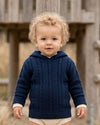 Carriage Boutique navy cable knit baby hooded sweater with cream cuffs worn by a toddler outdoors.