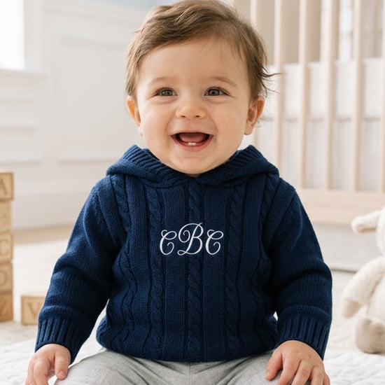 Carriage Boutique navy cable knit baby sweater with hood and white monogram embroidery on chest worn by a smiling baby