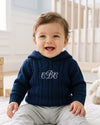 Carriage Boutique navy cable knit baby sweater with hood and white monogram embroidery on chest worn by a smiling baby