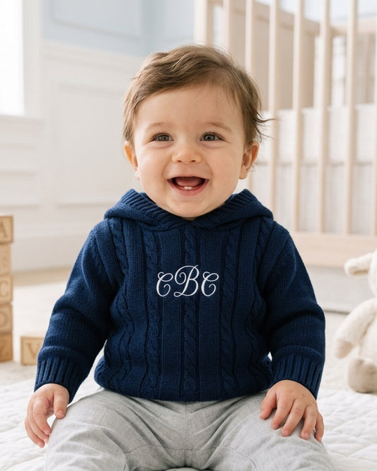 Carriage Boutique navy cable knit baby sweater with hood and white monogram embroidery on chest worn by a smiling baby